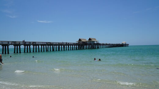 Ein langer Holzsteg führt über türkisfarbenes Meer hinaus, Menschen spazieren darauf, während andere im flachen Wasser am Strand baden. Der Himmel ist klar und blau, die Szene wirkt ruhig und einladend.