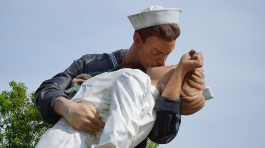 Große Außenskulptur eines Matrosen, der eine Krankenschwester in den Armen hält und küsst. Der Matrose trägt eine dunkle Uniform mit weißer Mütze, die Krankenschwester ein weißes Kleid und weiße Schuhe. Die Figuren stehen vor blauem Himmel und grüner Vegetation.