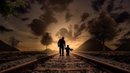 Vater und Sohn laufen auf verlassenen Bahnschienen Hand in Hand dem Sonnenuntergang entgegen.